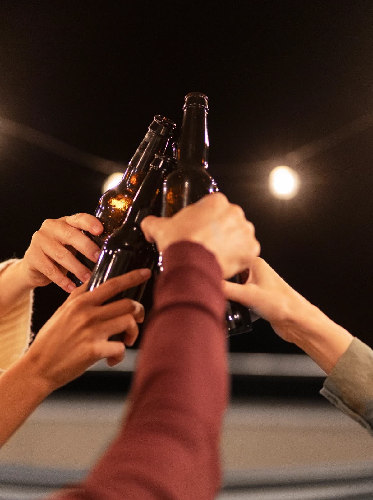 Four bottles meeting in a toast under a string light, late at night.