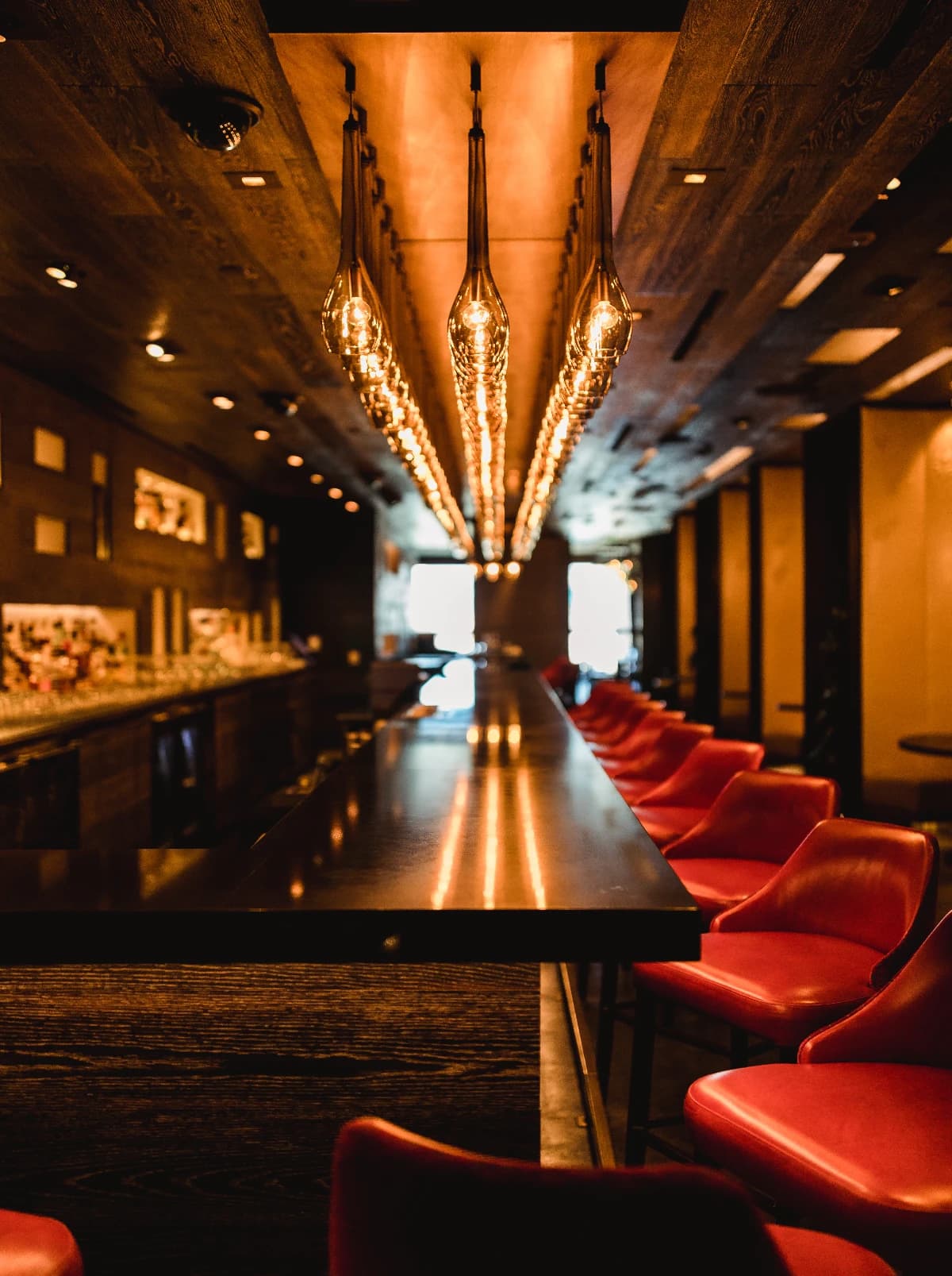 An empty bar counter lit by hanging bulbs, red leather stools waiting along it.