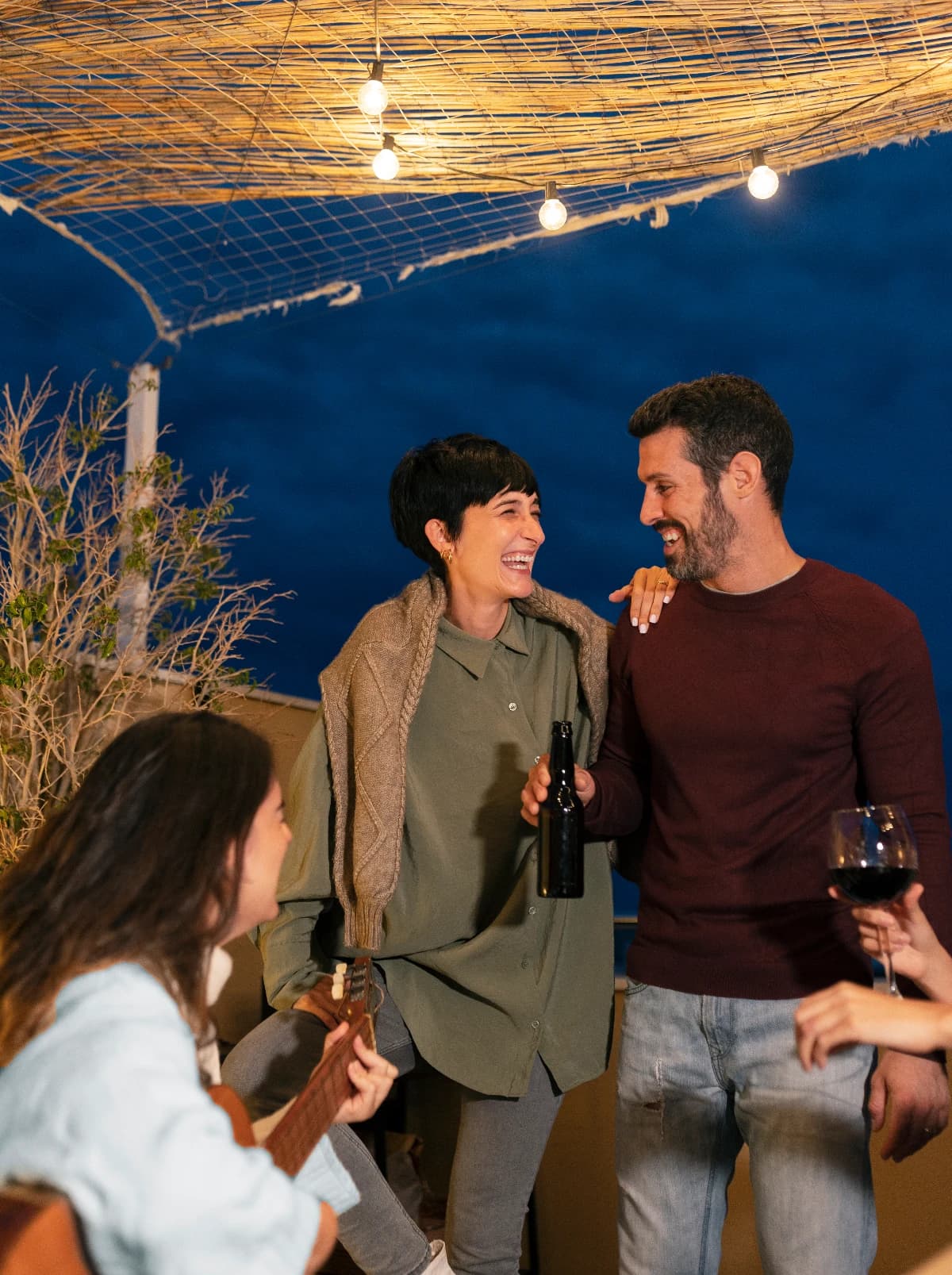Four people laughing together on a rooftop at dusk, string lights overhead.